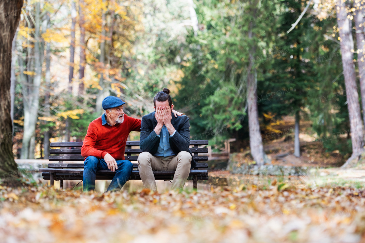 Senior father comforting his sad and frustrated adult son on bench in nature.