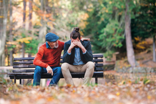 Senior father comforting his sad and frustrated adult son on bench in nature.