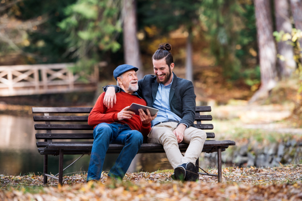 Senior father and his young son sitting on bench by lake in nature, using tablet.