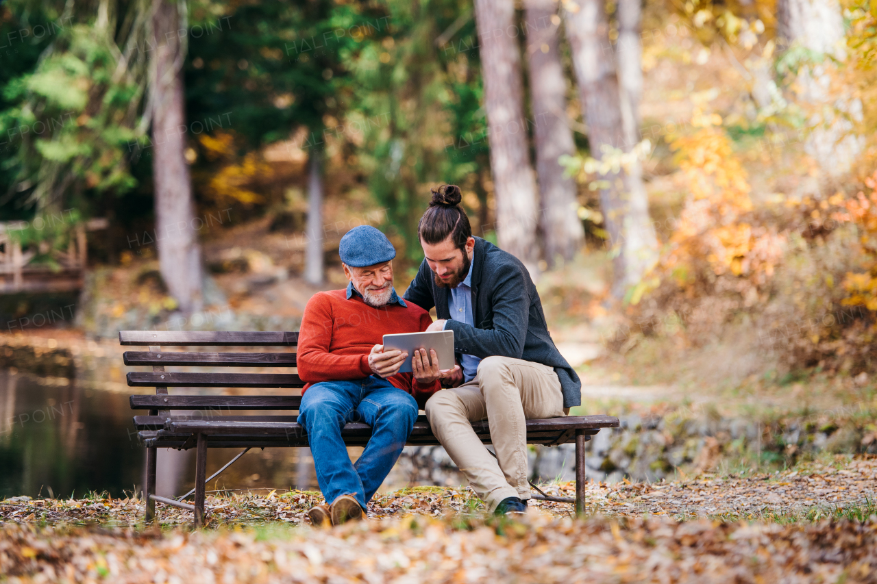 A senior father and his son sitting on bench in nature, using tablet.