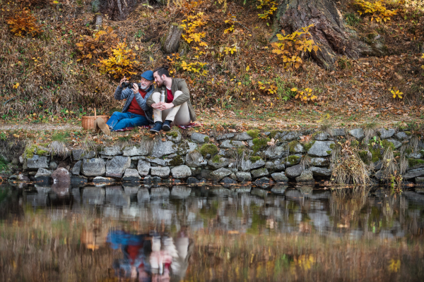 Senior father and his son with binoculars and picnic basket sitting by lake in nature.