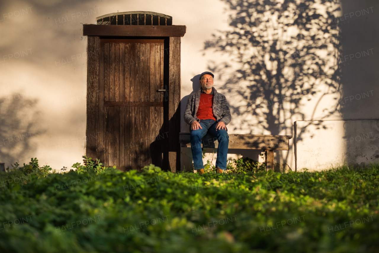 A lonely senior man sitting on bench in front of old house, eyes closed.