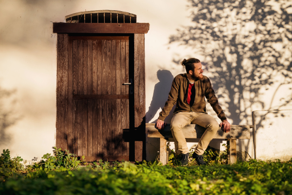 Front view of lonely young man sitting on bench in front of old house.