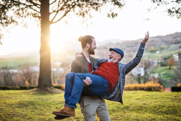 A young man carrying his senior father in arms in nature, having fun.