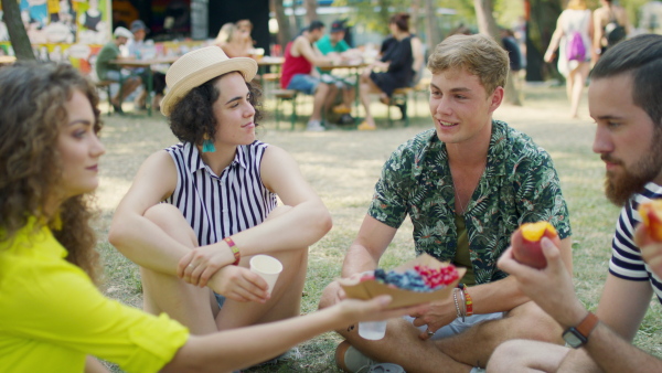 Group of young cheerful friends at summer festival, sitting on the ground and eating.