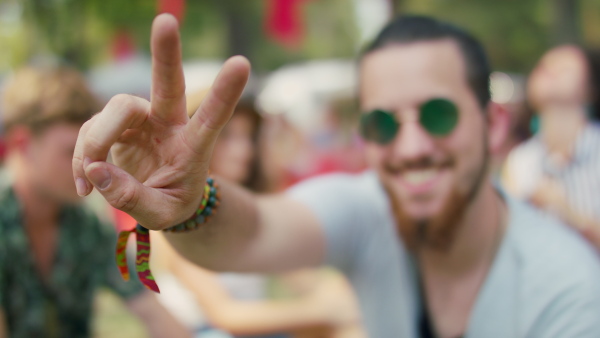 A front view of young man sitting at summer festivaland showing victory sign.