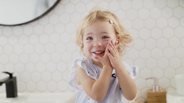 A front view of happy small girl indoors in bathroom, putting cream on face.