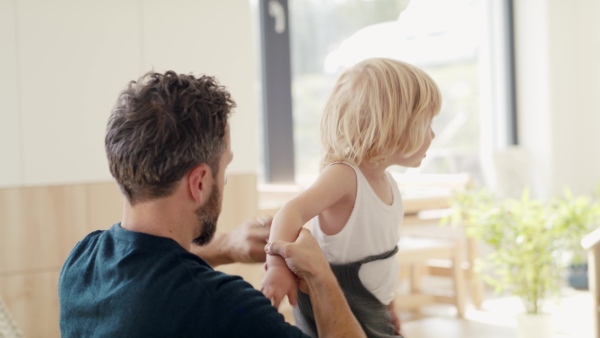 A young father helping small son to get dressed indoors in bedroom.