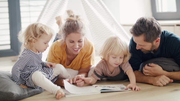 Front view of young family with two small children indoors in bedroom reading a book.