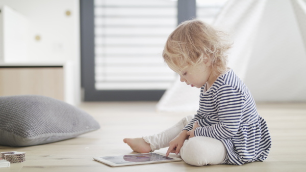 A cute small girl sitting indoors on the floor in bedroom, using tablet.