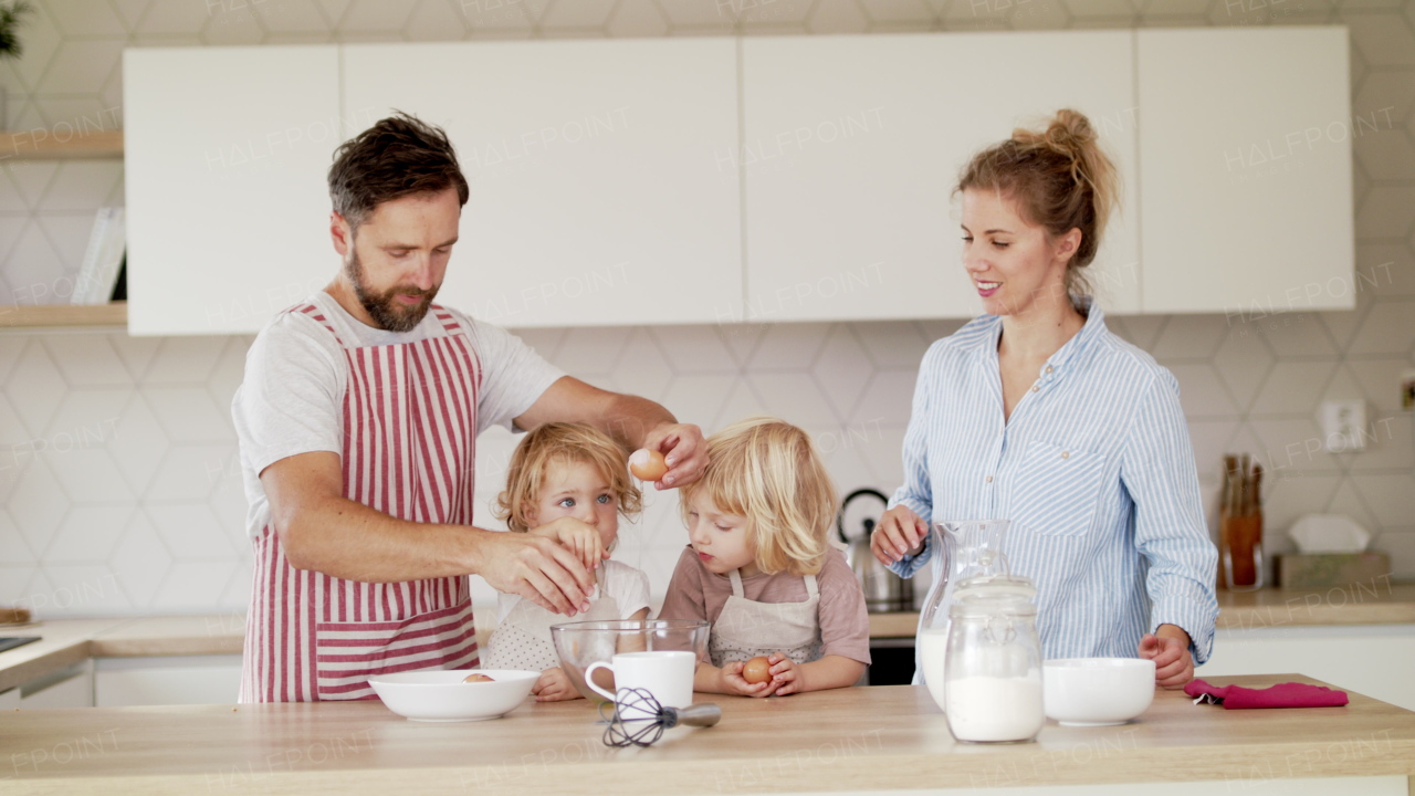 Front view of young family with two small children indoors in kitchen, breaking eggs when cooking.