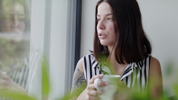 A happy young woman sitting by window indoors at home drinking tea or coffee.