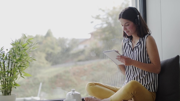 A young woman with headphones sitting by window indoors at home writing and listening to music.