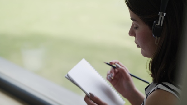 A young woman with headphones sitting by window indoors at home writing and listening to music.