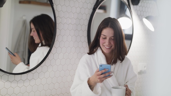 A young woman with coffee standing indoors in bathroom at home using smartphone.