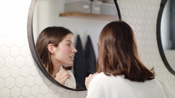 A happy young woman standing indoors in bathroom at home looking in mirror.