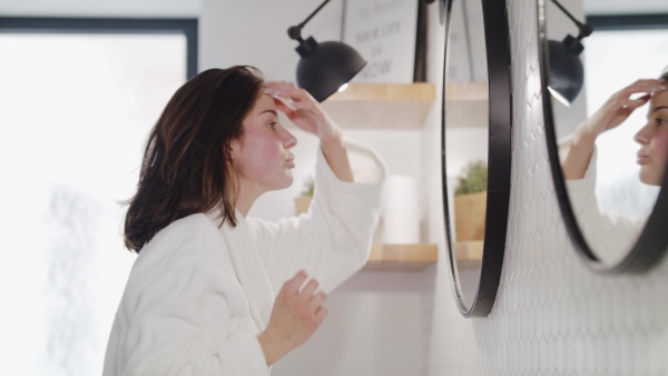 A happy young woman standing indoors in bathroom at home looking in mirror.