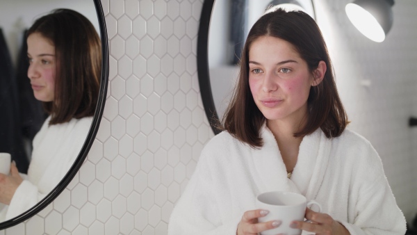 A young woman standing indoors in bathroom at home drinking tea or coffee.