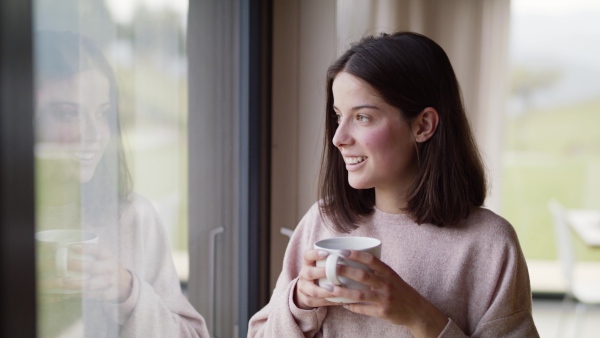 A happy young woman standing by window indoors at home drinking tea or coffee.