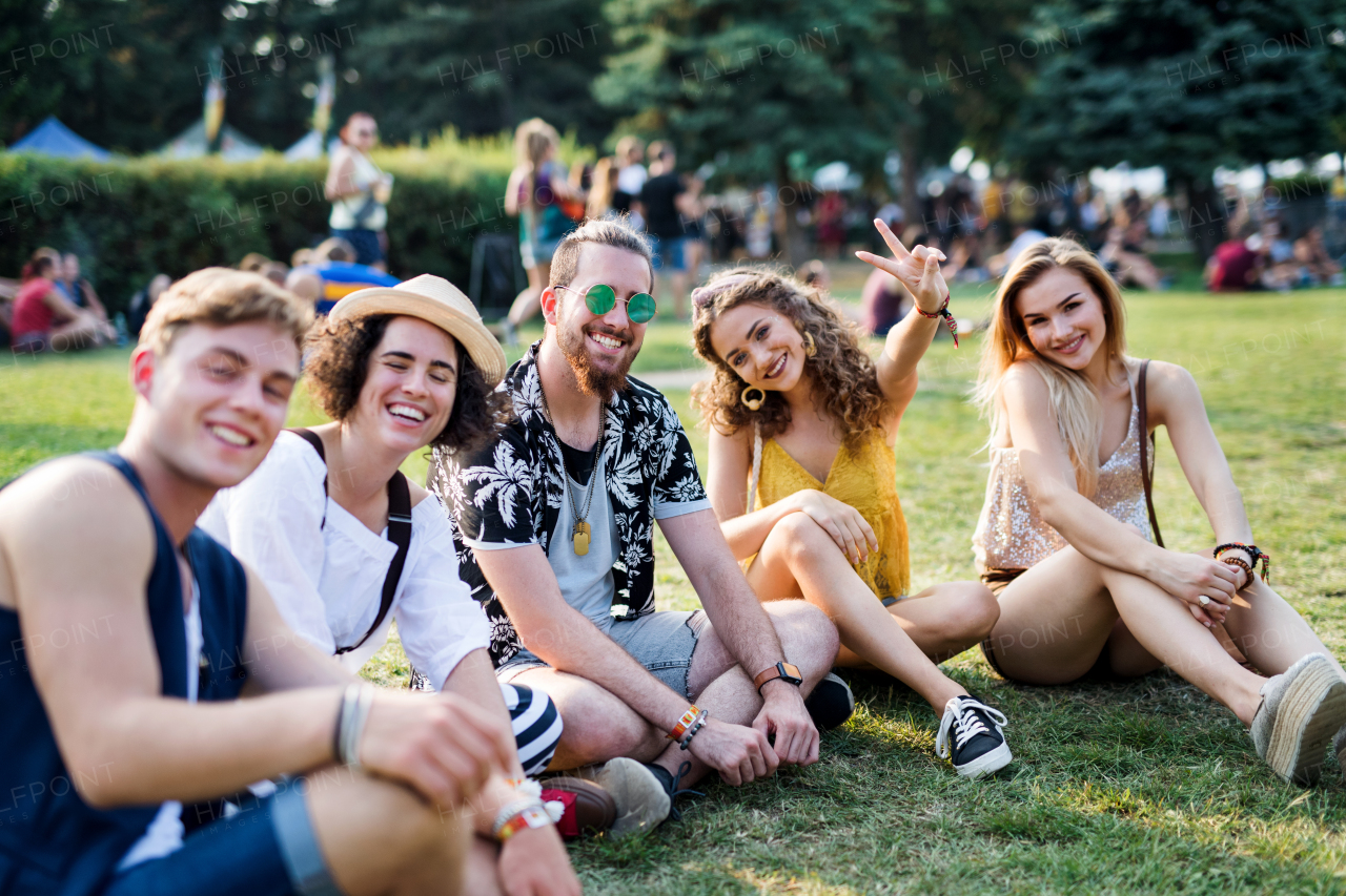 Group of cheerful young friends sitting on ground at summer festival, looking at camera.