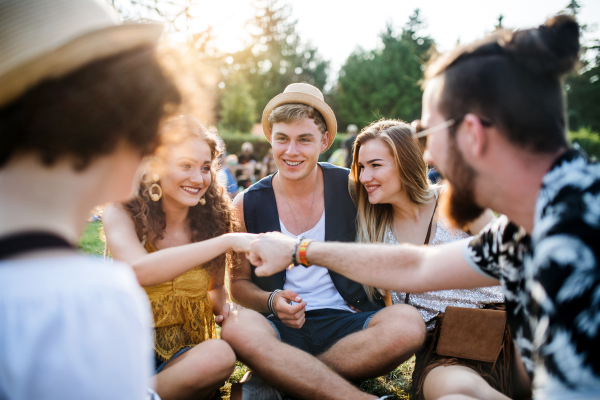 Group of cheerful young friends sitting on ground at summer festival, giving fist bump.