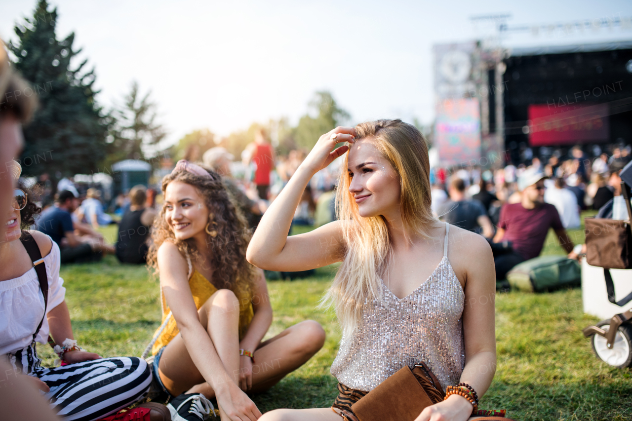 Group of cheerful young female friends sitting on ground at summer festival, talking.