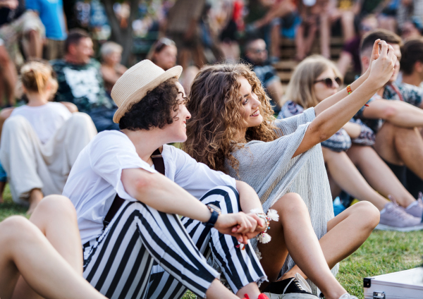 Group of cheerful young friends sitting on ground at summer festival, taking selfie.