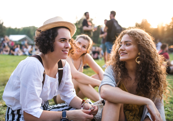 Group of cheerful young female friends sitting on ground at summer festival, talking.