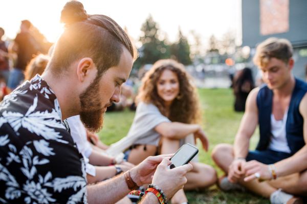 Group of cheerful young friends sitting on ground at summer festival, looking at camera.