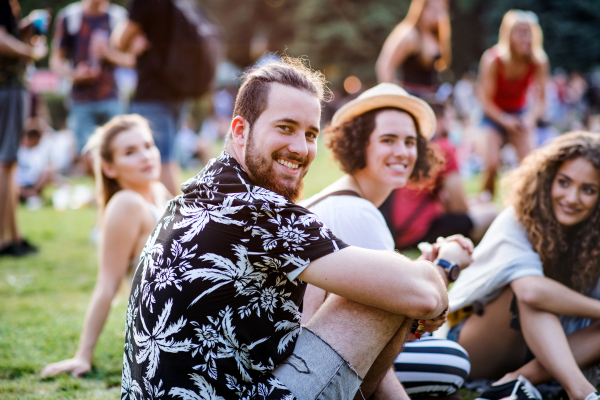 Group of cheerful young friends sitting on ground at summer festival, looking at camera.