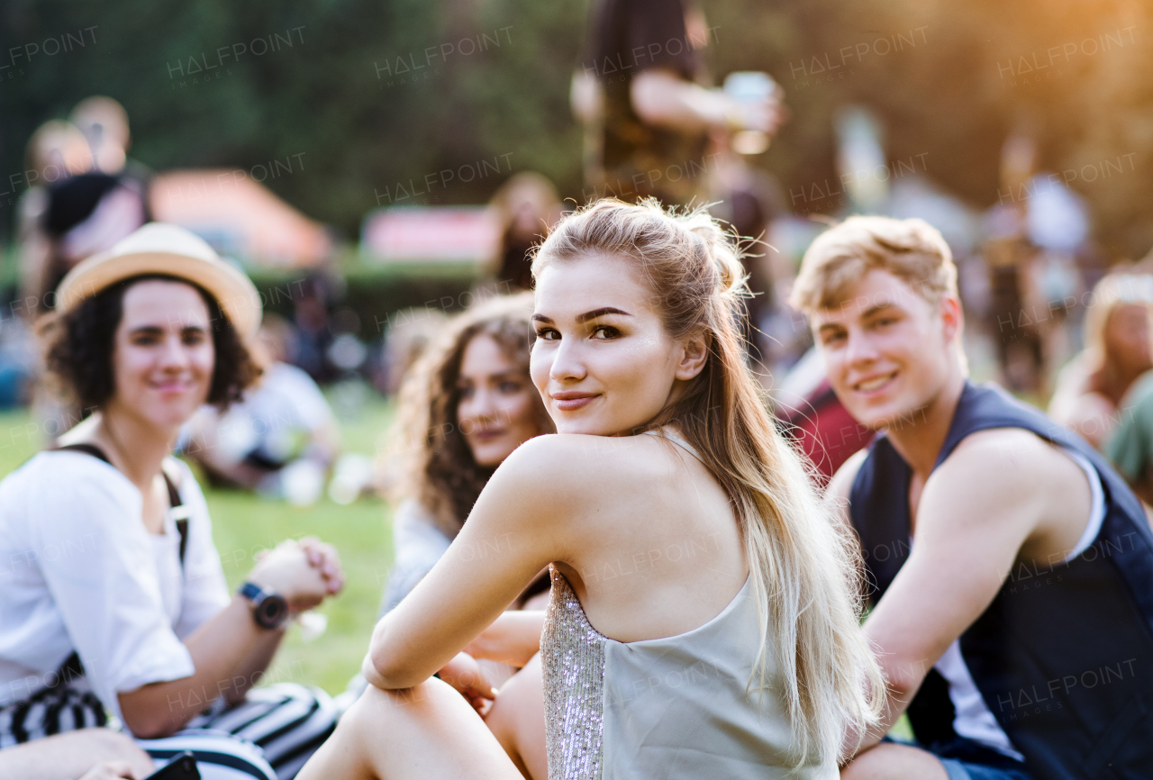 Group of cheerful young friends sitting on ground at summer festival, looking at camera.