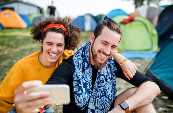 Front view of young couple at summer festival or camping holiday, taking selfie with smartphone.