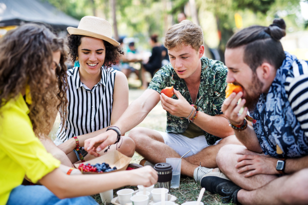 Group of young cheerful friends at summer festival, sitting on the ground and eating.