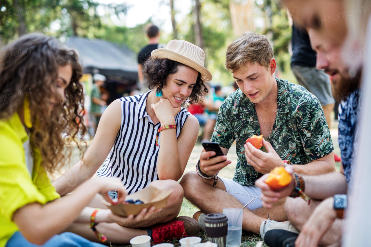 Group of young cheerful friends at summer festival, sitting on the ground and eating.