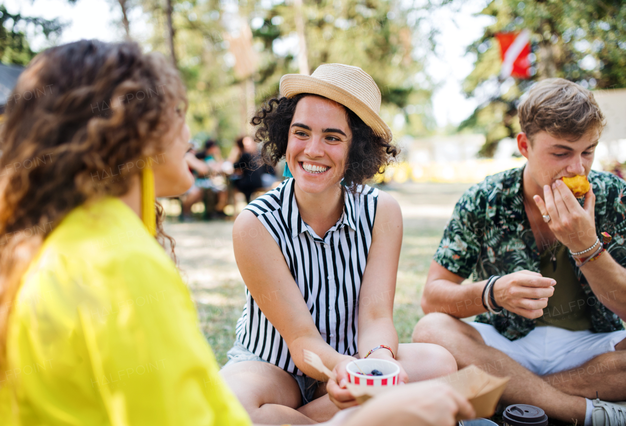 Group of young cheerful friends at summer festival, sitting on the ground and eating.