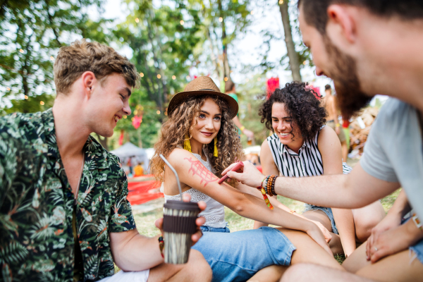 Group of cheerful young friends sitting on ground at summer festival, drawing on body.