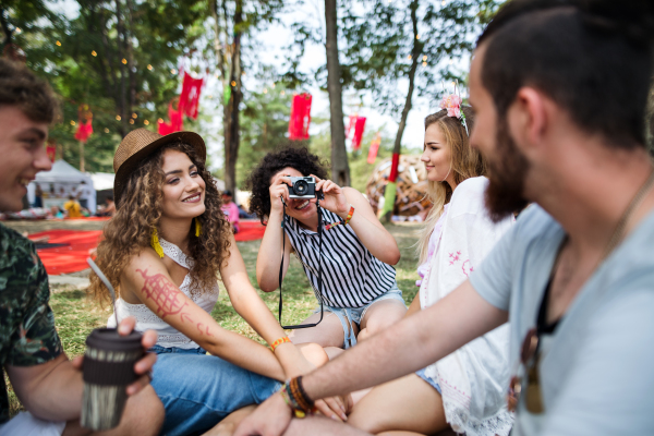 Group of young friends sitting on ground at summer festival, taking photo with camera.