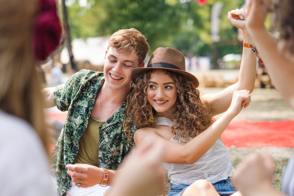 Group of cheerful young friends sitting on ground at summer festival, dancing.