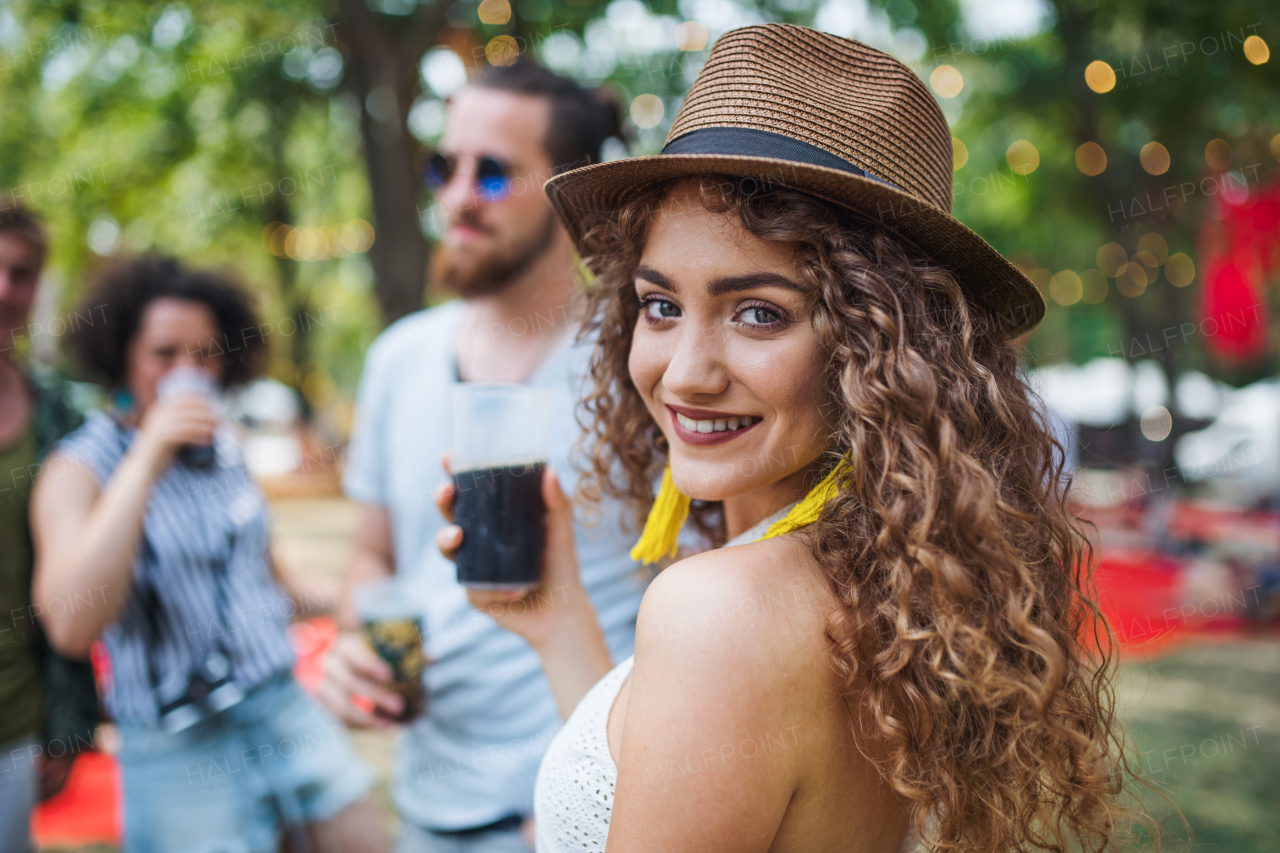 Beautiful young girl with drink and friends standing at summer festival.