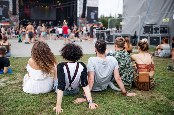 Rear view of group of unrecognizable young friends at summer festival, sitting on ground.