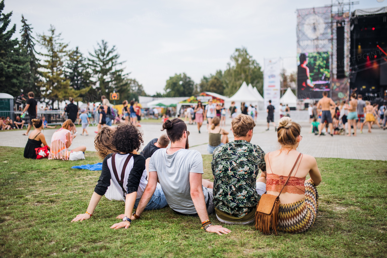 Rear view of group of unrecognizable young friends sitting at summer festival.