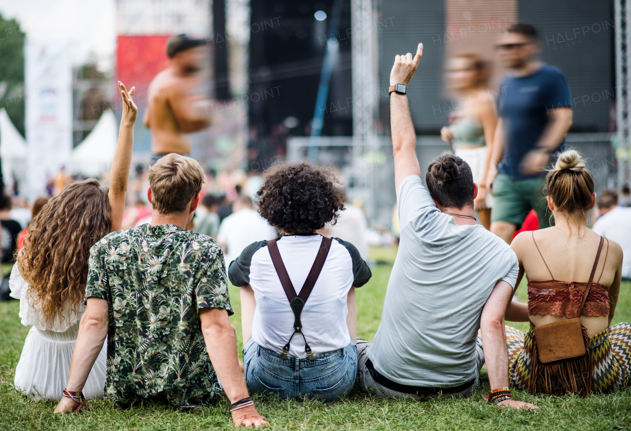 Rear view of group of unrecognizable young friends at summer festival, sitting on ground