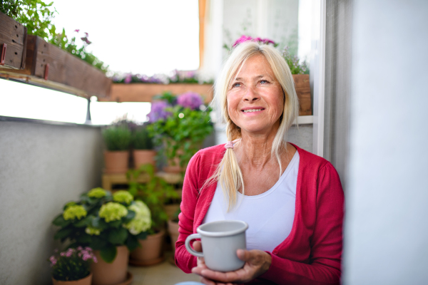 Happy senior woman with coffee on balcony in summer, relaxing among potted flowering plants.