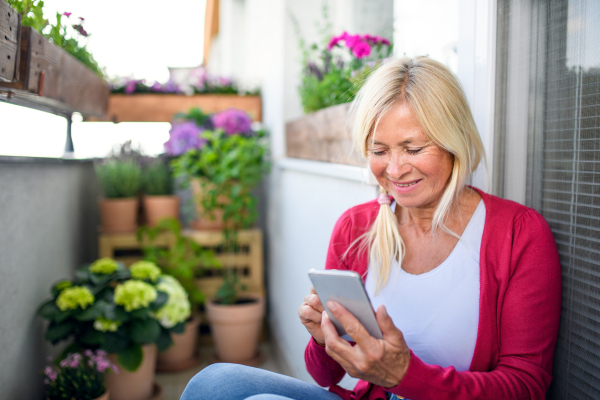 Happy senior woman relaxing on balcony in summer, using smartphone.