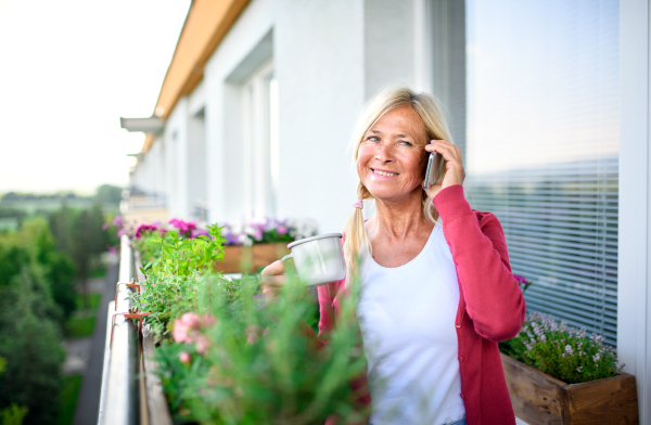 Happy senior woman with coffee relaxing on balcony in summer, using smartphone.