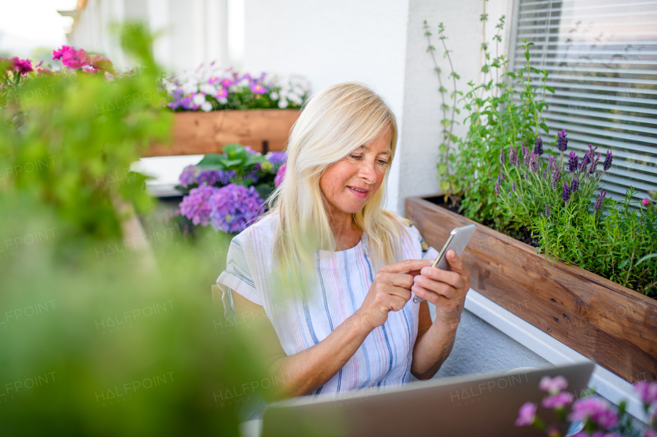 Active senior woman with laptop and smartphone working on balcony in summer, home office.