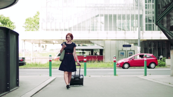 Young woman commuter with suitcase walking outdoors in city, using smartphone.