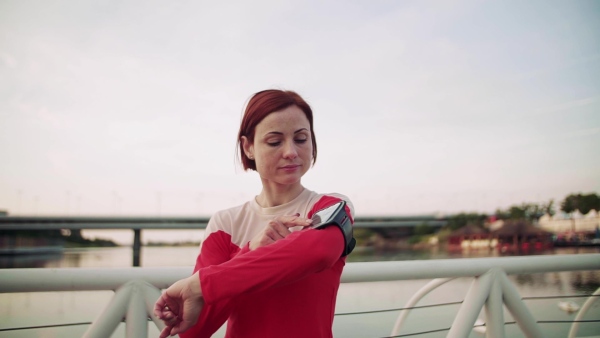 A young woman resting after doing exercise on bridge outdoors in city, using smartphone. Slow motion.