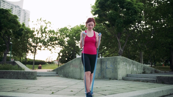 Full length portrait of young woman doing exercise outdoors in city with elastic bands. Slow motion.