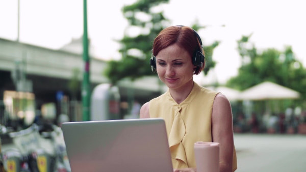 A young businesswoman with laptop and headphones sitting in cafe outdoors in city, working.